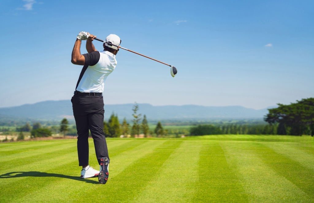 Golfer swinging club on a sunny golf course with scenic background.